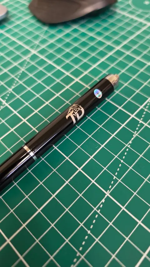 Custom laser engraved black metal pen with precise tiger monogram on green grid cutting mat for personalized branding.