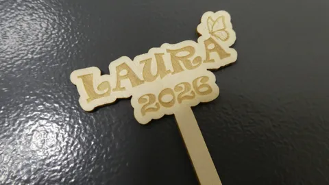 Custom laser-cut wooden cupcake topper featuring precise engraving of the name Laura 2026 with a butterfly detail.