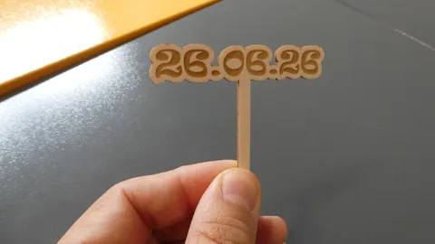 Close-up of a hand holding a tiny laser-cut wooden topper precisely engraved with the date 26.06.26.